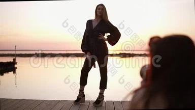 美丽的高大模特，穿着黑色连衣裙和凉鞋，站在大海前，为相机摆好姿势